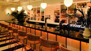 Zona de bar con varios taburetes, una barra repleta de botellas y copas, e iluminación colgante.