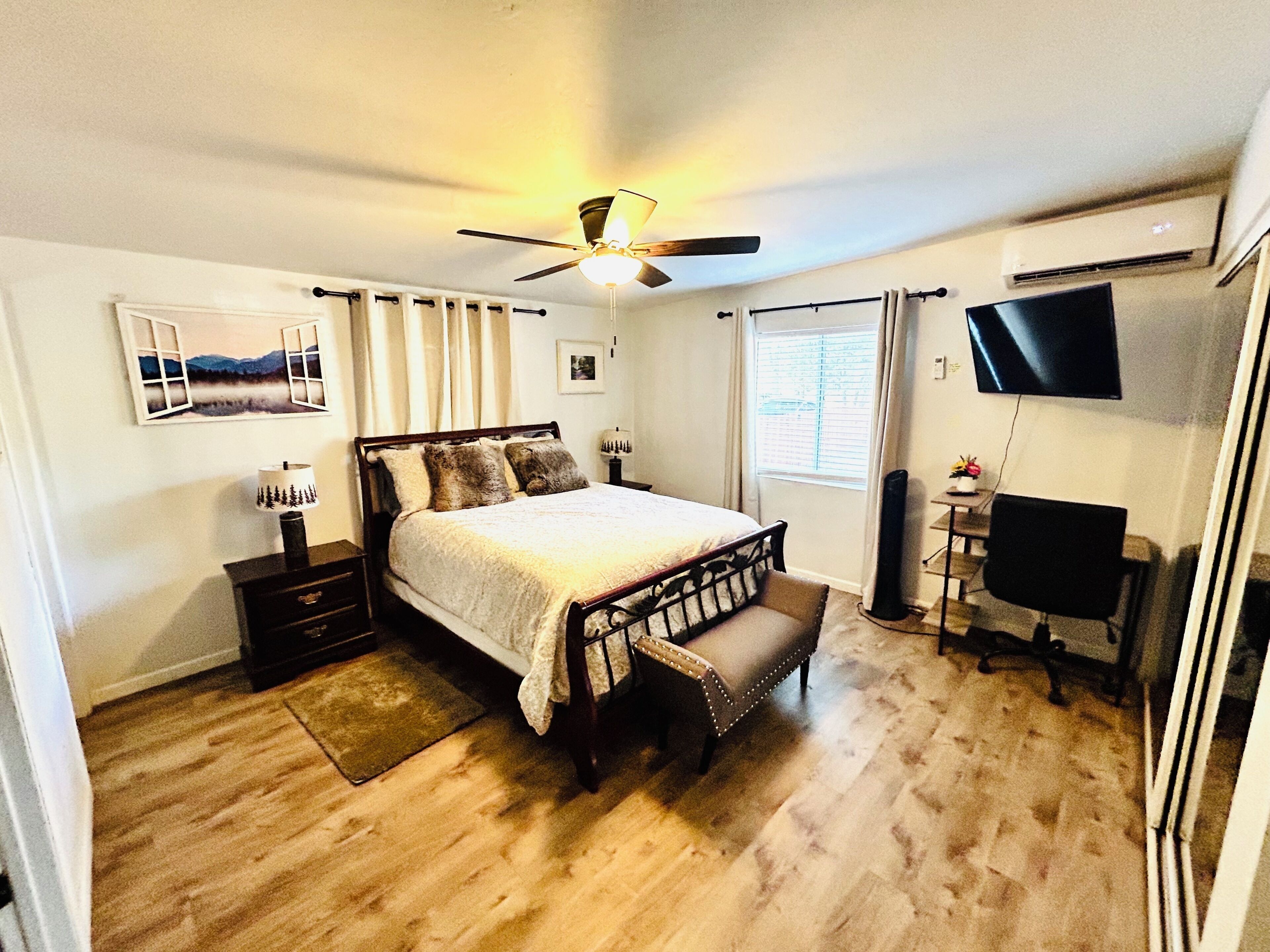A bedroom with a bed, bedside tables, a desk, a chair, a ceiling fan, a TV, and a window with curtains.