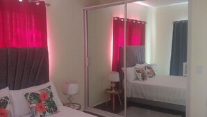 Habitació d'hotel amb un armari amb mirall, un llit amb almoines florals, una taula de nit amb una làmpada i cortines vermelles.