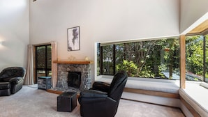 A modern living room with a stone fireplace, leather chairs, and large windows offering a view of greenery.