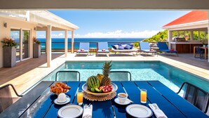 Un espace repas aménagé au bord de la piscine, avec une table dressée pour le petit-déjeuner, offrant une vue sur la piscine et l’océan.