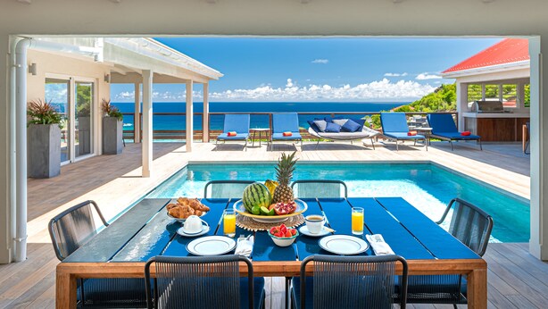 Un espace repas aménagé au bord de la piscine, avec une table dressée pour le petit-déjeuner, offrant une vue sur un océan d’un bleu limpide.