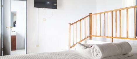 Un dormitorio con cama, una escalera de madera, un televisor montado en la pared y una puerta que da al baño.