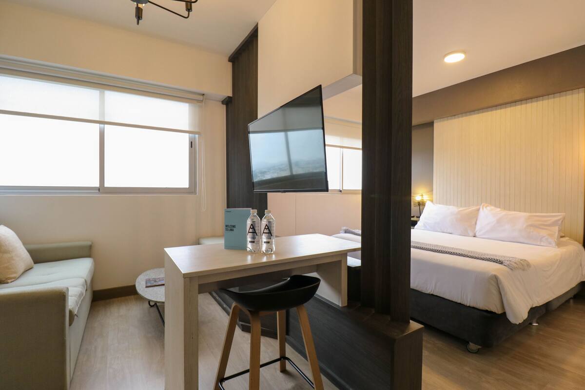 A hotel room with a bed, a desk with a chair, a small table, and a window with blinds.