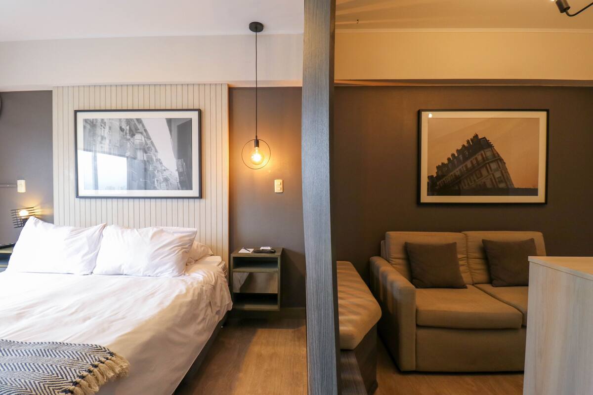 A modern hotel room with a large bed, a sofa, a desk, and two framed pictures on the walls.