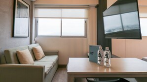Een hotelkamer met een bank, een tafel met waterflesjes, een televisie en een raam met luxaflex.