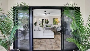 A modern living room with a white sofa, a coffee table, and a ceiling fan. There are potted plants on either side of the sliding glass door. - Tranquil Palm Cove Retreat with Heated Pool, Lush Gardens & Steps to the Beach (Palm Cove)