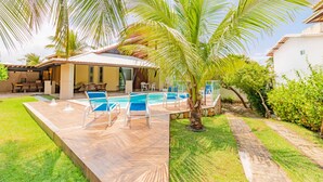 Uma área de piscina com espreguiçadeiras, um espaço de estar coberto e uma palmeira.