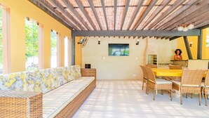 Uma área de estar coberta ao ar livre, com sofá e mesa de jantar, rodeada por cadeiras de vime.