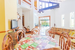 Uma sala de jantar com uma mesa redonda, cadeiras de vime e toalha de mesa colorida. Há uma televisão sobre um suporte à esquerda.