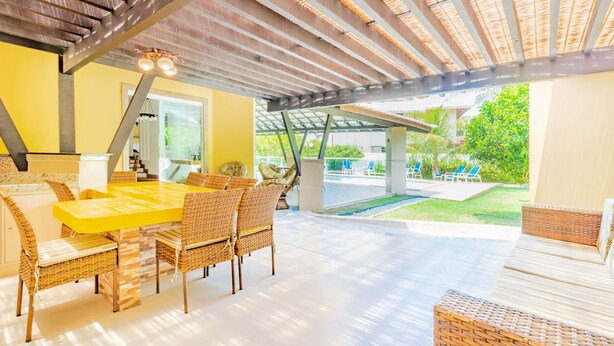 Uma área de refeições ao ar livre coberta, com mesa amarela e cadeiras de vime, uma pérgola acima e vista para uma piscina e vegetação.