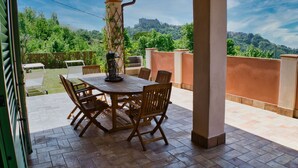 Eine Terrasse im Freien mit Tisch und Stühlen, einem Pergola und Blick auf einen Hügel mit Bäumen und einem schlossartigen Bau im Hintergrund.