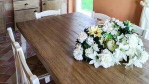 Un comedor con una mesa de madera, sillas blancas, un centro de mesa floral, una cocina con horno blanco y una alfombra estampada en la pared.