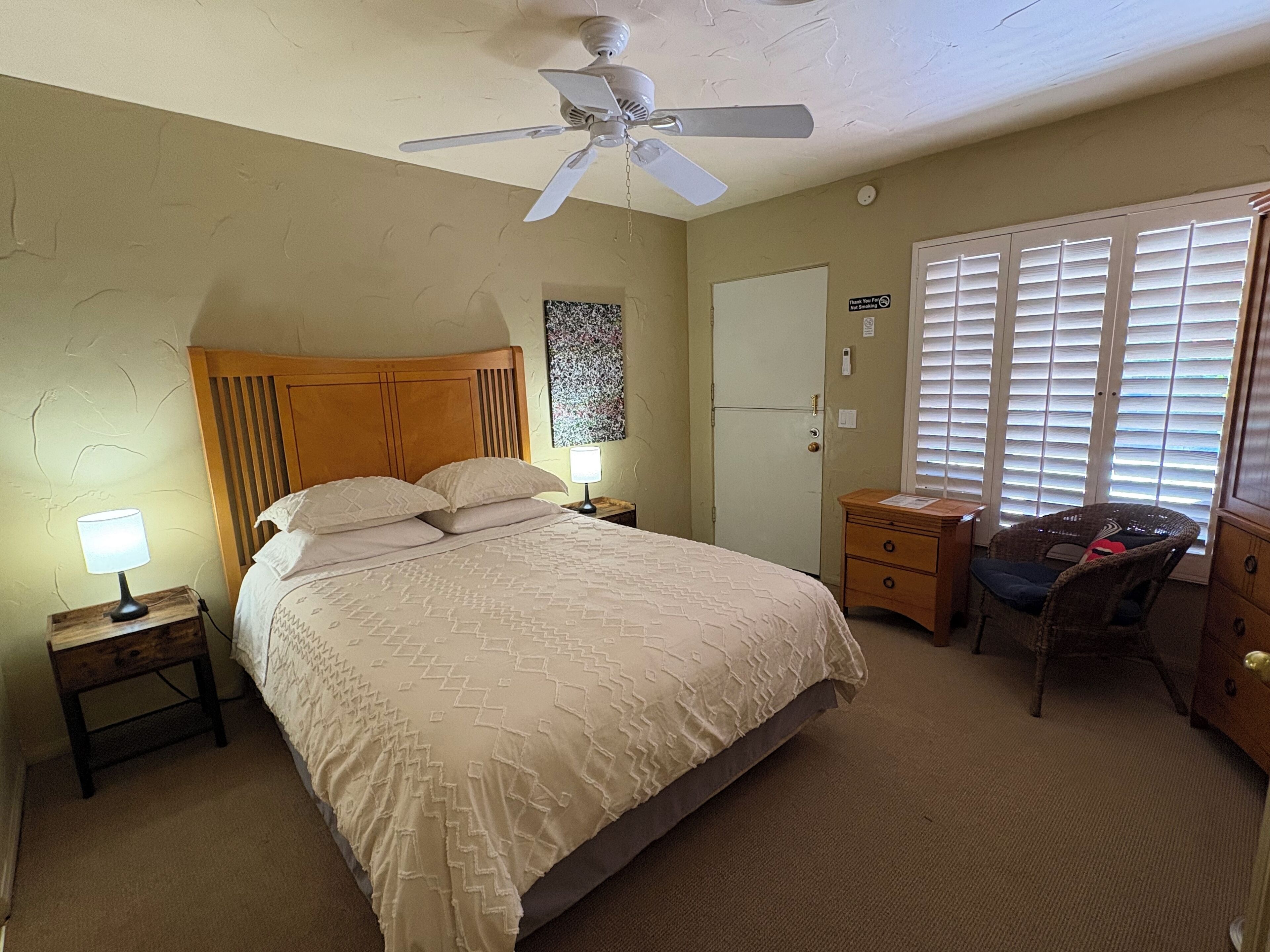 A bedroom with a bed, a nightstand, a desk, a chair, a ceiling fan, a door, and a window with blinds.