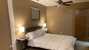 Ein ordentlich bezogenes Bett mit weißer Bettwäsche, zwei Nachttischlampen, einem Deckenventilator und einem Fenster mit Jalousien.