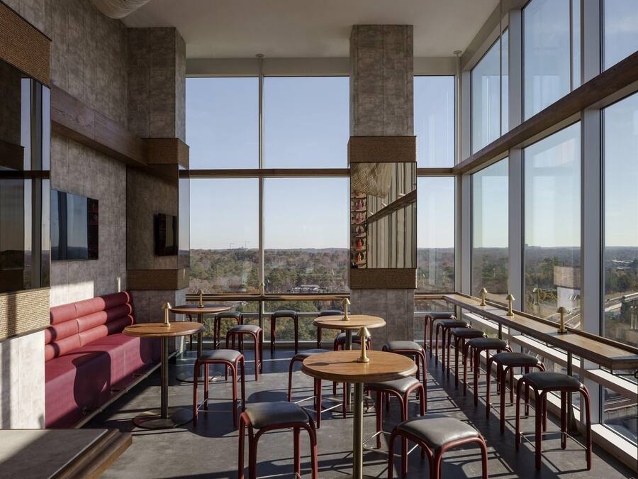 A modern interior with a red sofa, round tables, and bar stools, large windows offering a city view.