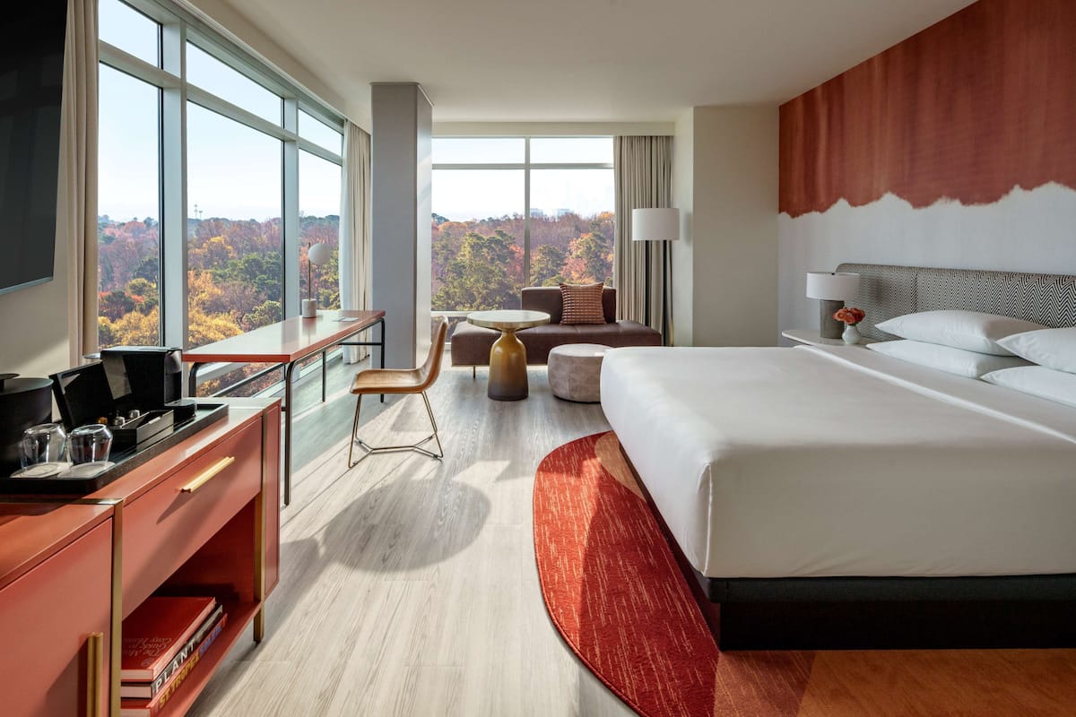A hotel room with a large bed, a desk with a laptop, a chair, and a view of autumn foliage through large windows.