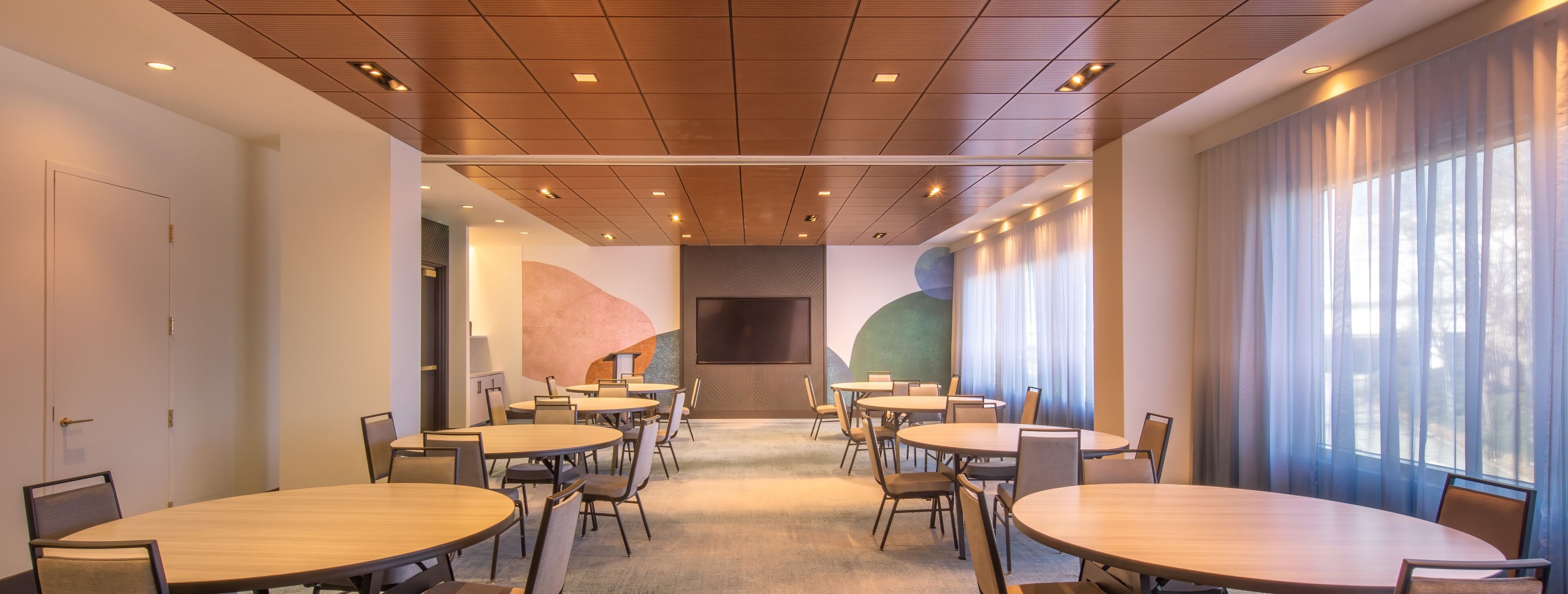 A conference room with multiple round tables and chairs, a large screen, and a view through large windows.