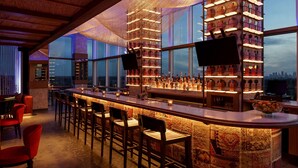 Eine Bar mit einer hohen Theke, verschiedenen Sitzgelegenheiten und Blick auf die Stadt durch große Fenster.