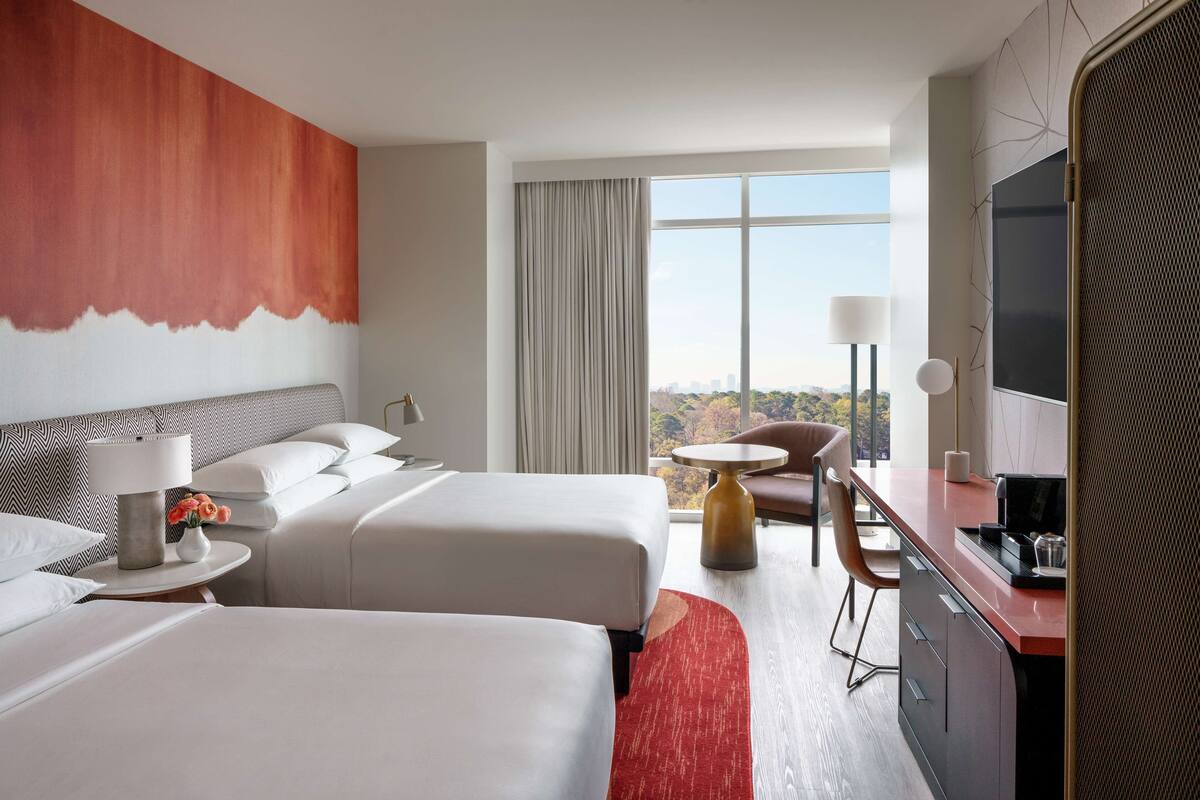 A hotel room with two beds, a desk, a chair, a TV, and a large window with a view.