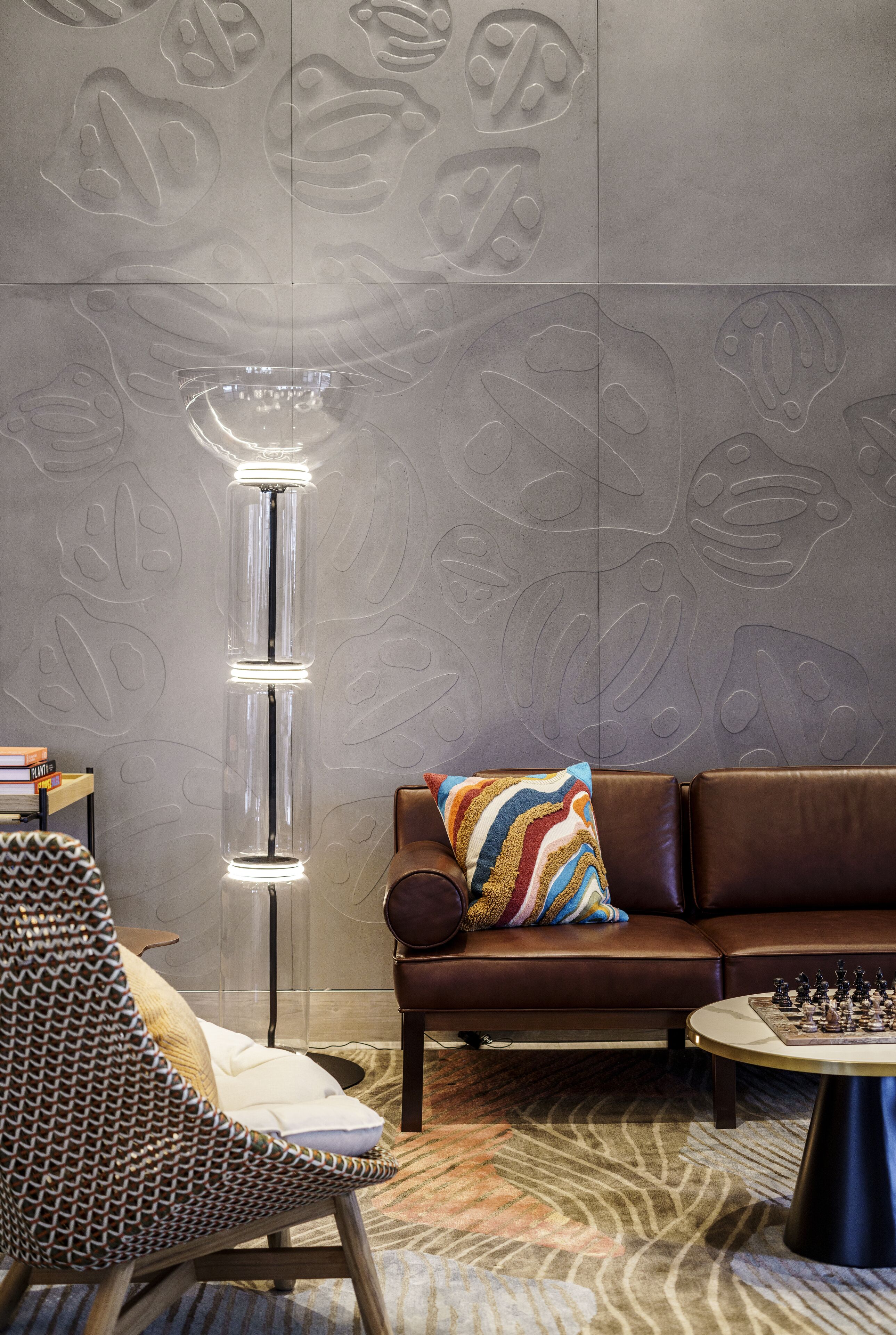 A modern living room with a patterned wall, a tall floor lamp, a brown leather sofa, a patterned armchair, and a chess table.