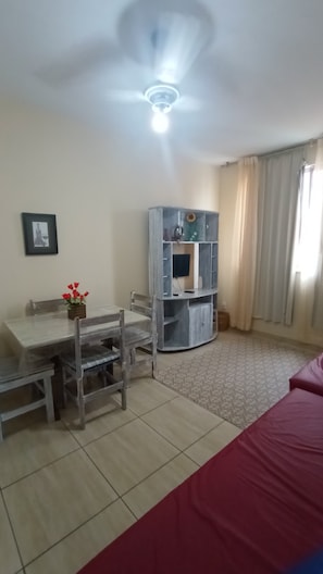 A room with a dining table, chairs, a red bed, a ceiling fan, and a TV on the wall. - Near Praia do Forte—Wi-Fi and parking available! Quiet neighborhood! Less than 100 meters from the beach! (Cabo Frio)