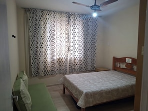 A bedroom with a bed, a wooden headboard, a green bench, a ceiling fan, and patterned curtains. - Near Praia do Forte—Wi-Fi and parking available! Quiet neighborhood! Less than 100 meters from the beach! (Cabo Frio)