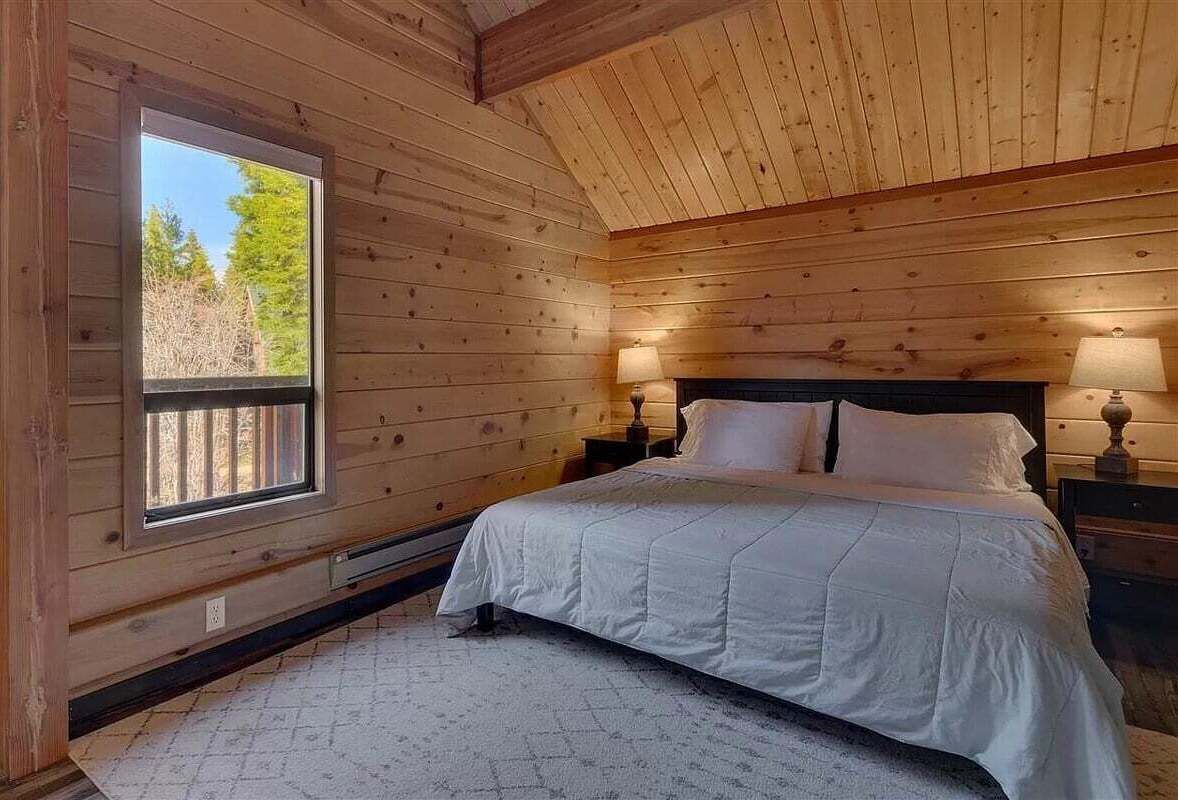 A wooden cabin bedroom with a bed, two bedside lamps, a window, and a view of trees.