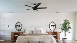 Quarto com cama, duas mesas de cabeceira, um banco, ventilador de teto e planta em vaso.