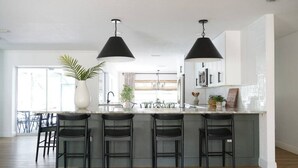 Cozinha moderna com ilha central, banquetas pretas, iluminação pendente e uma grande janela que proporciona luz natural.