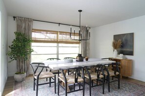 Uma sala de jantar com uma mesa branca, cadeiras pretas, um lustre, um vaso de planta e um quadro na parede.
