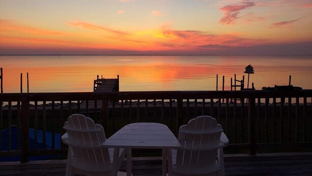 A wooden deck with two white Adirondack chairs and a table, overlooking a calm body of water during sunset.