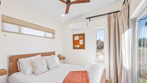 Een slaapkamer met een bed, een plafondventilator, een raam met gordijnen en uitzicht op de omgeving.