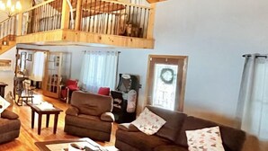 A spacious living area with a high ceiling, wooden beams, a brown sofa, a coffee table, and a patterned rug. - Private Adirondack Getaway. Close to hiking trails, fishing, public beach. (Clemons)