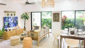 Un salón amplio con sofá de madera, mesa y sillas de comedor, televisor y un gran ventanal con vistas a zonas verdes.
