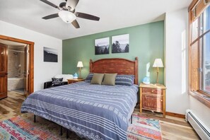 Une chambre à coucher comprenant un lit, deux tables de chevet, un ventilateur de plafond, une fenêtre et une porte donnant sur une salle de bain.