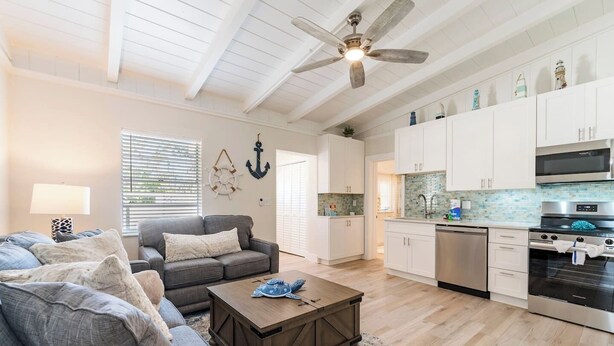 A spacious living room with a grey sofa, a wooden coffee table, and a ceiling fan. Adjacent is a kitchen with white cabinets, a stainless steel oven, and a microwave. - Cute Home with pool, spa, mini golf walk to beach! (Holmes Beach)