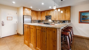 A kitchen with wooden cabinets, a stainless steel refrigerator, a microwave, and a sink. There are bar stools and pendant lights. - Lodge at Mtn Village B147 | Ski-In/Out | Valley View | Large Balcony | Washer/Dryer | Pool | Hot Tub (Park City)