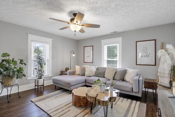 A living room with a sectional sofa, a ceiling fan, and a large window with blinds. - Stylish Small-Town Retreat - Near Lancaster (Baltimore)