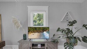 Un televisor sobre un soporte de madera, con una escena de playa, una ventana con persianas y una planta a la derecha.