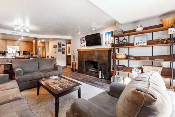 A spacious living room with a fireplace, a television mounted on the wall, and a kitchen in the background.