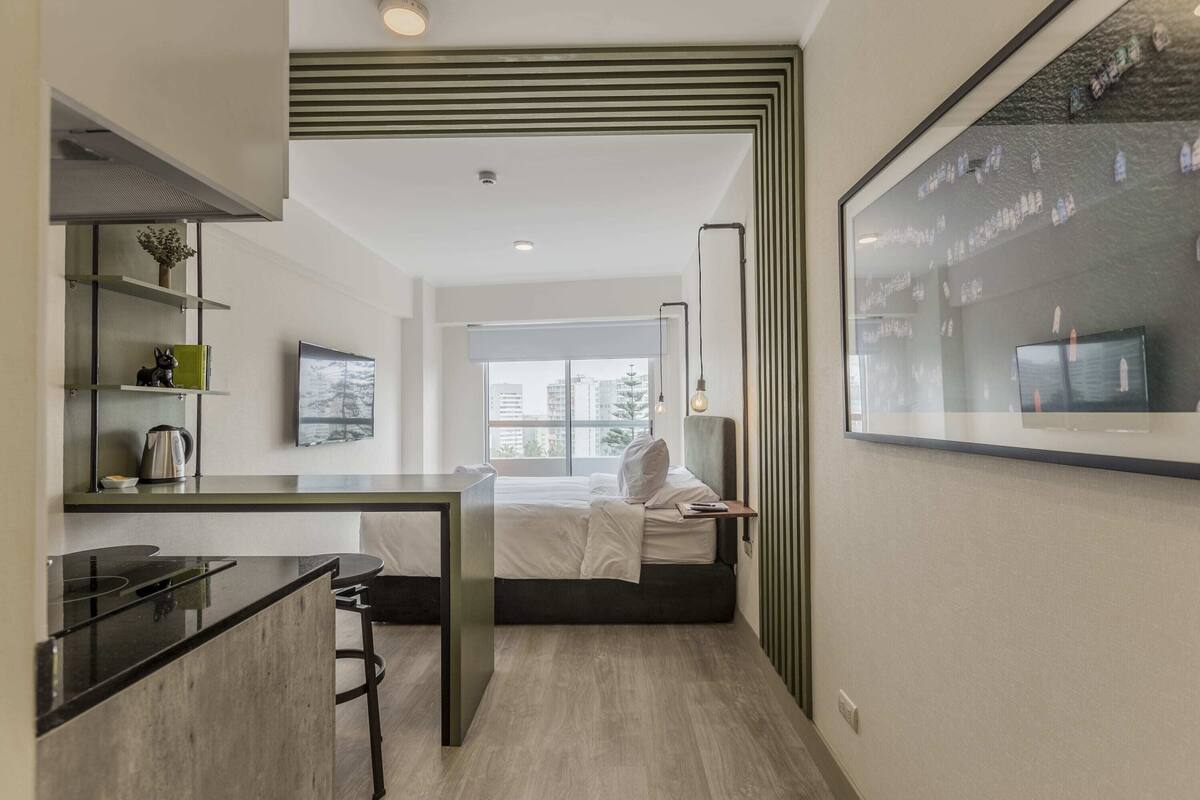 A modern hotel room with a kitchenette, a bed with white bedding, a large window, and a wall-mounted TV.