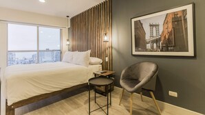 Ein modernes Hotelzimmer mit einem großen Bett, einem kleinen runden Tisch, einem grauen Sessel, Blick auf die Stadt und einem gerahmten Bild an der Wand.