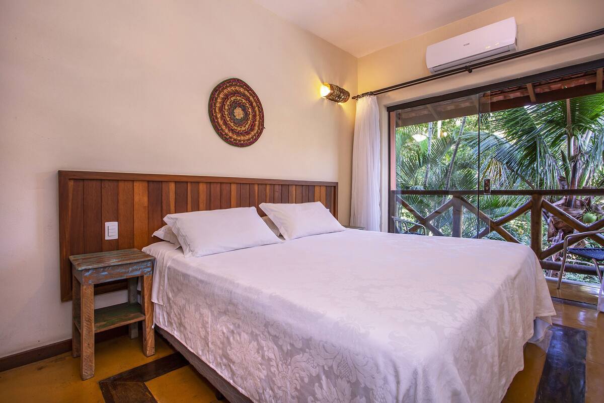 A bedroom with a wooden headboard, a white bedspread, a small wooden nightstand, and a large window with a view of greenery.
