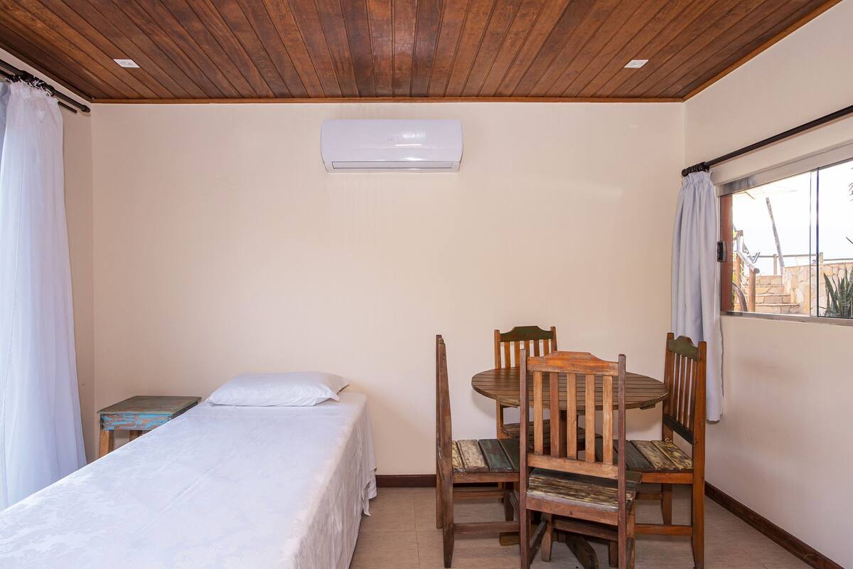 A bedroom with a bed, a wooden table with chairs, a window with curtains, and an air conditioning unit on the wall.