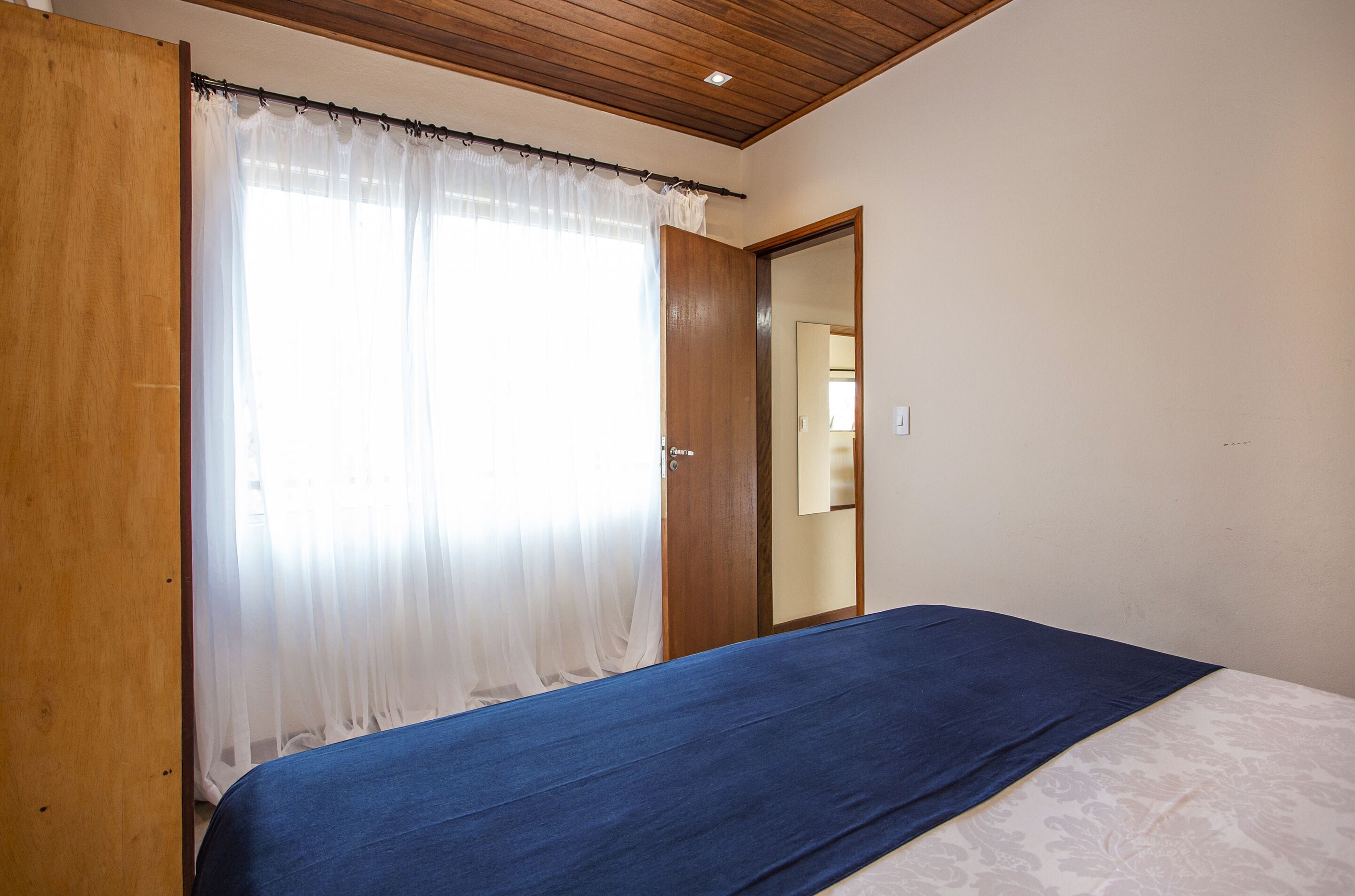 A bedroom with a bed, a wooden door, and a window with sheer curtains.