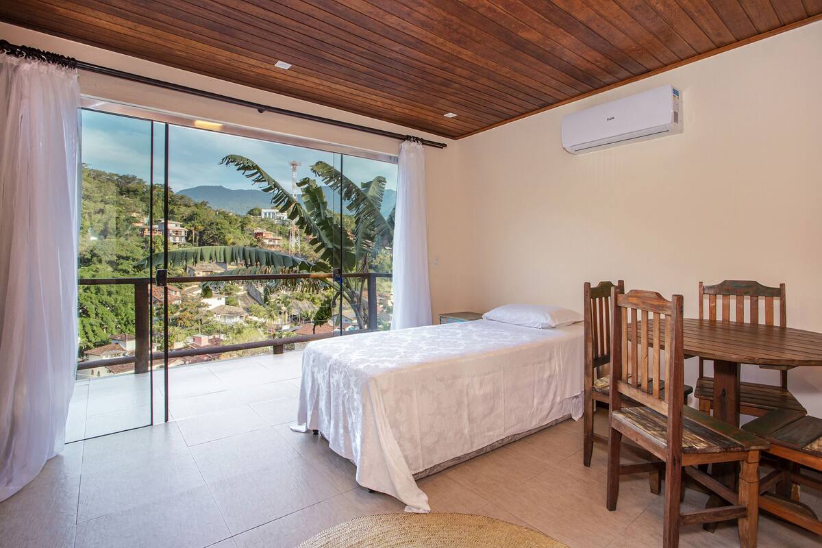 A bedroom with a large window, a bed, a wooden table, and a chair.