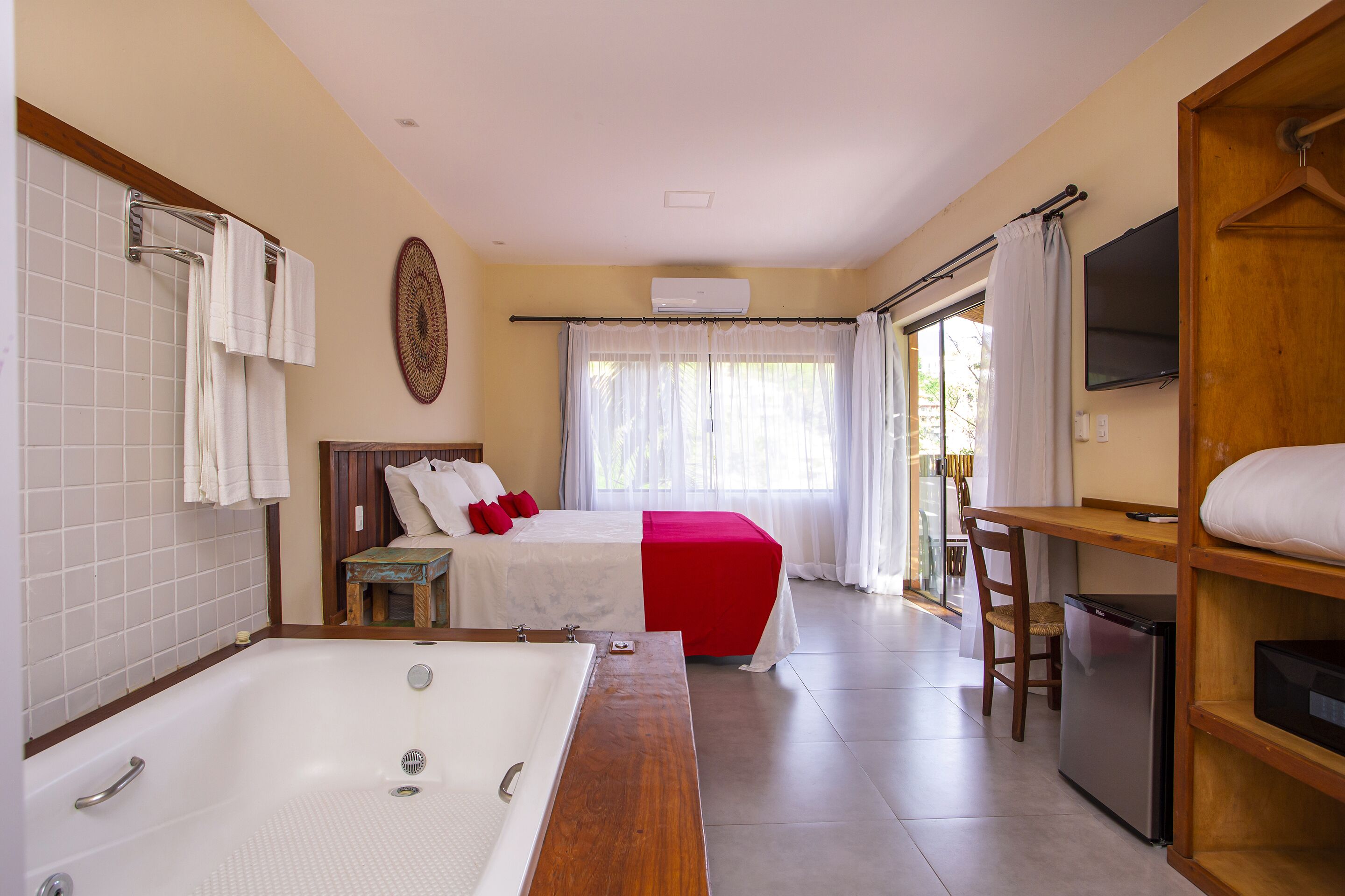 A hotel room with a large bathtub, a bed with red bedding, a small table, a chair, and a TV on a wooden shelf.