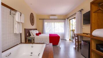A hotel room with a large bathtub, a bed with red bedding, a small table, a chair, and a TV on a wooden shelf.