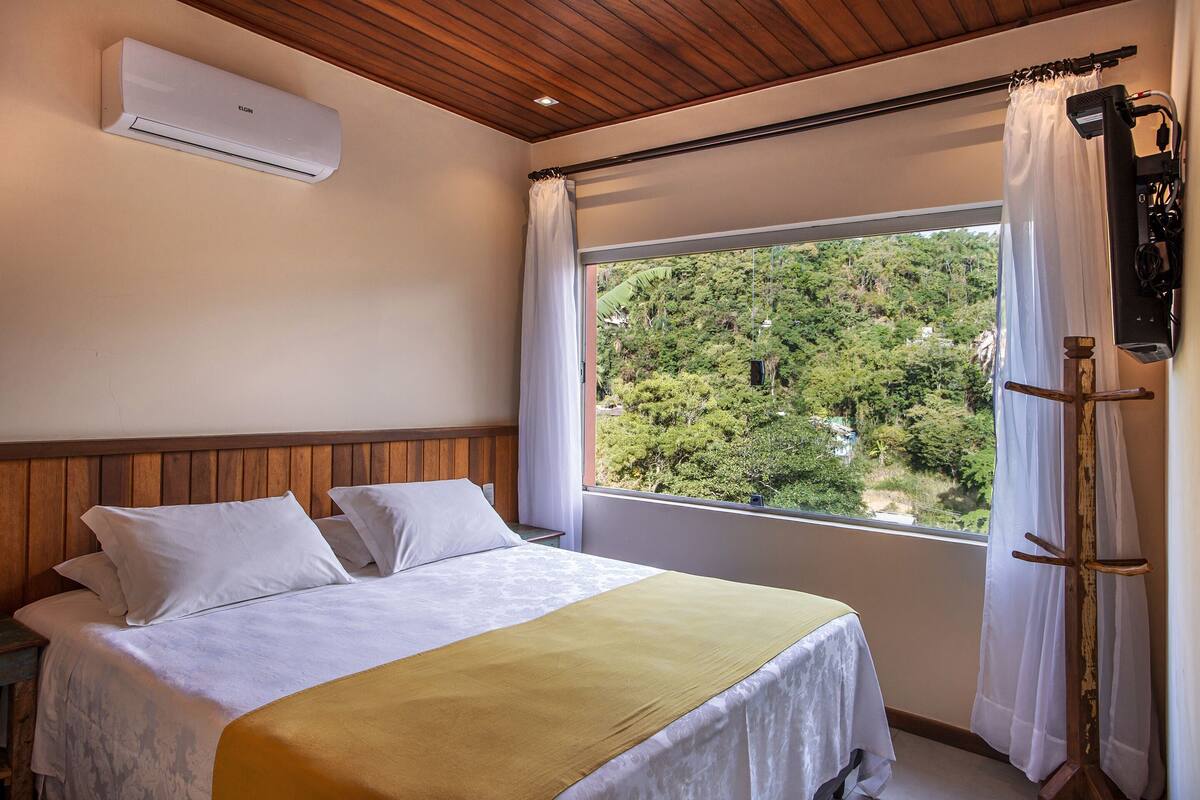 A bedroom with a bed, a window with curtains, an air conditioning unit, and a wooden ceiling.
